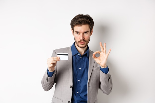 Cliente do sexo masculino feliz em terno de negócio, mostrando cartão de crédito de plástico e sinal de ok, sorrindo satisfeito