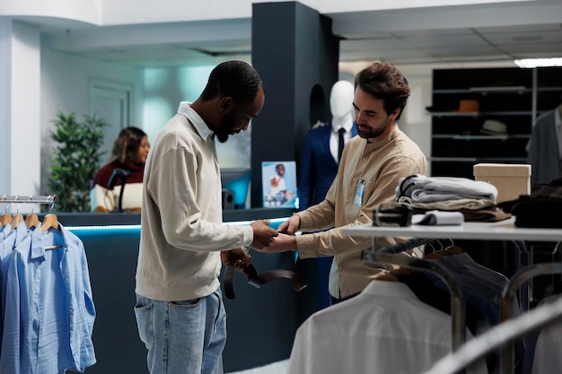 Cliente e trabalhador de loja de roupas examinando a qualidade e o estilo do cinto de couro. Homem afro-americano segurando acessório e pedindo conselhos ao assistente de boutique enquanto compra roupas masculinas