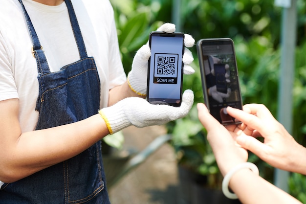 Cliente escaneando el código qr en la pantalla del teléfono inteligente en manos del trabajador del centro de jardinería para pagar la compra