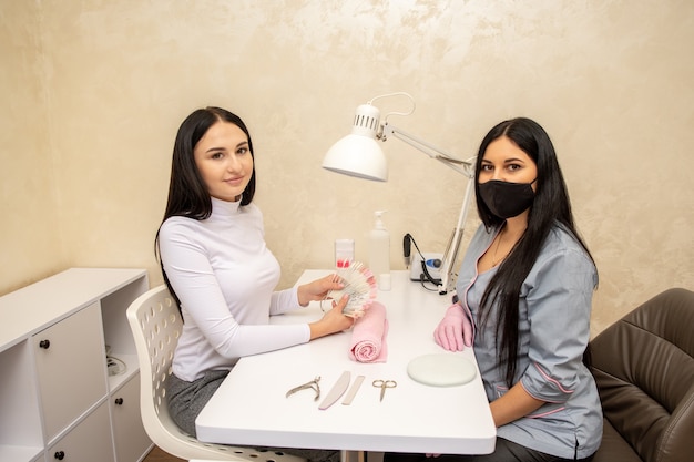 Foto cliente escolhendo a cor do esmalte para as unhas com a manicure no salão de beleza. mestre da manicure usando máscara