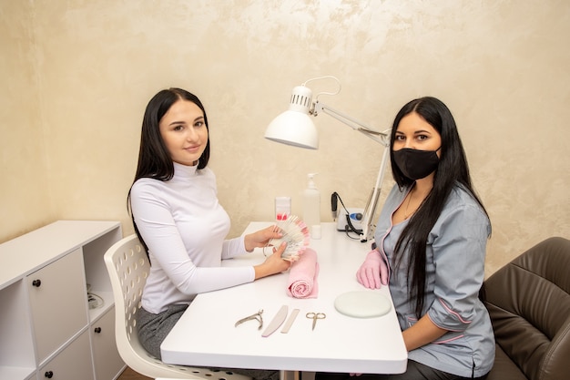 Cliente escolhendo a cor do esmalte para as unhas com a manicure no salão de beleza. Mestre da manicure usando máscara