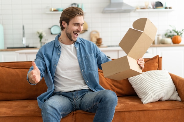 El cliente feliz desembalaje de la caja de cartón recibe el paquete de entrega de correo postal abierto. Sonriente joven comprador satisfecho desembalaje paquete desembalaje orden de envío compra de productos sentarse en el sofá en casa