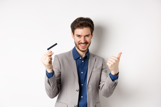 Cliente feliz en traje de negocios mostrando tarjeta de crédito plástica y pulgar hacia arriba, sonriendo satisfecho