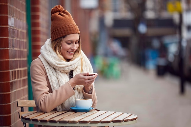 Cliente femenino que envía un mensaje de texto sentado afuera de una cafetería en la concurrida calle principal de la ciudad en otoño