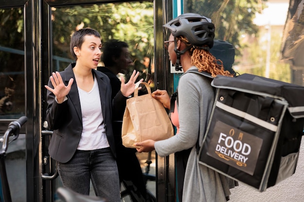 El cliente insatisfecho se niega a tomar un pedido de comida incorrecto del servicio de mensajería del servicio de entrega, cliente enojado. Mujer dando comida para llevar a un empleado furioso e impulsivo cerca del edificio de la empresa