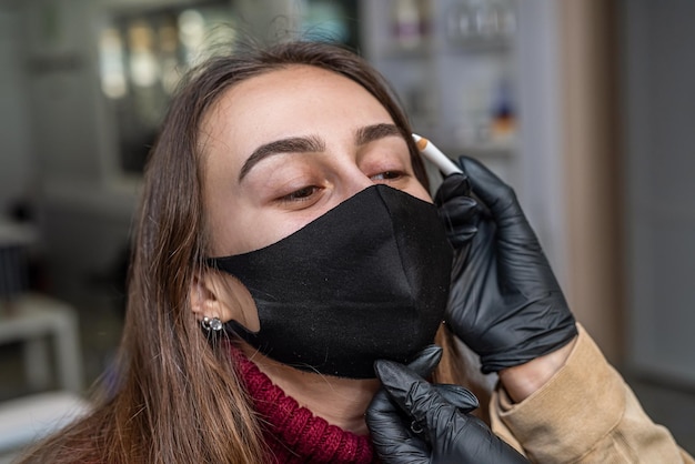 Cliente jovem e bonita em um mestre de máscara preta desenha uma pálpebra com um lápis branco