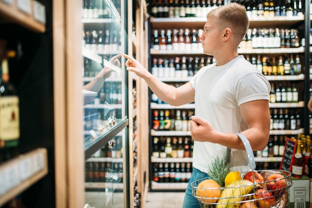 Cliente masculino con cesta eligiendo cerveza en el supermercado. Compras en la tienda de alimentos, sección de alcohol en el fondo