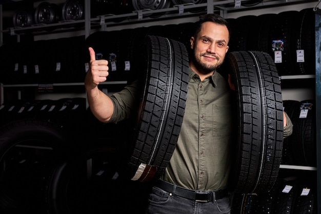 El cliente masculino está satisfecho con la compra de un neumático en el garaje de reparación de automóviles, soporte que sostiene el caucho para el automóvil en las manos, en la tienda de la tienda de automóviles