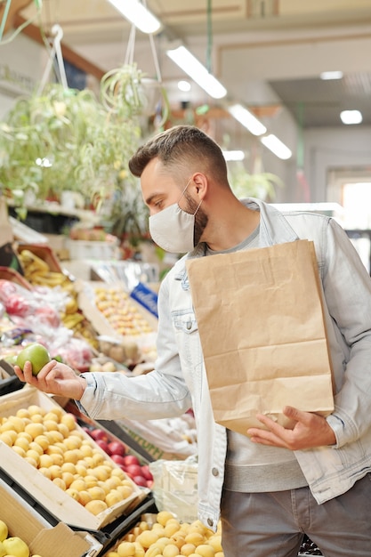 Cliente masculino joven en máscara de tela de pie en el mostrador de alimentos y sosteniendo una bolsa de papel mientras compra manzanas en el mercado de agricultores