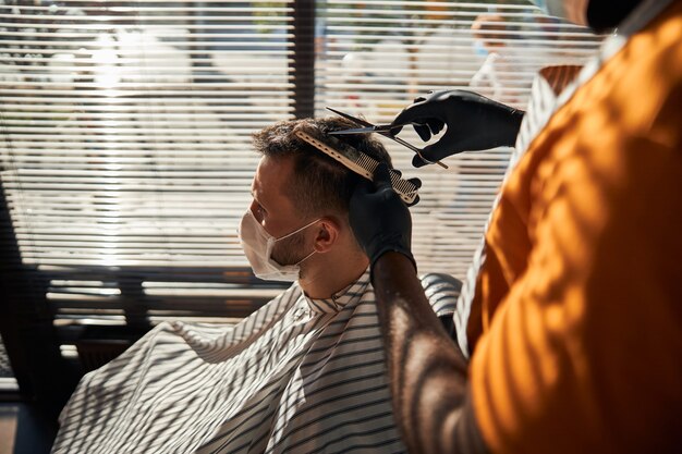 Cliente masculino en máscara médica cortándose el pelo en peluquería