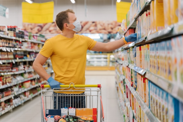 Cliente masculino posa no hipermercado em máscara protetora médica e luvas de borracha, compra os produtos necessários