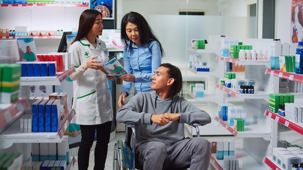 Cliente masculino en silla de ruedas y cuidador hablando con un farmacéutico, buscando comprar medicamentos en la farmacia. Persona con discapacidad crónica que recibe ayuda médica y vitaminas, comprando medicamentos.