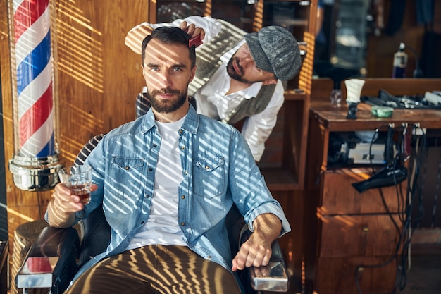 Foto cliente satisfecho de una peluquería sentado con un vaso de whisky mientras peluquero peina su cabello