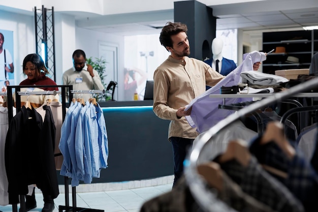 Cliente segurando roupas formais, examinando o tecido e o ajuste antes de fazer a compra na loja de roupas. Homem caucasiano escolhendo roupa e verificando o estilo da camisa na boutique do shopping