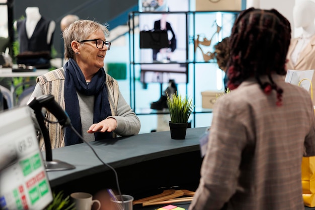 Cliente senior discutiendo con el trabajador de la sala de exposición en la caja registradora, pagando ropa de moda en el centro comercial. Mujer anciana adicta a las compras comprando mercancía elegante en boutique de moda