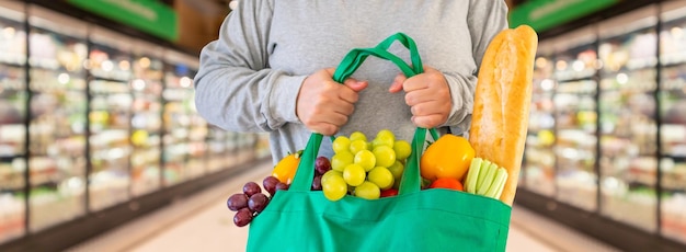 El cliente tiene una bolsa de compras verde reutilizable con frutas y verduras con fondo de pasillo de supermercado