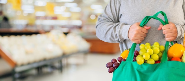 El cliente tiene una bolsa de compras verde reutilizable con frutas y verduras sobre el fondo del supermercado