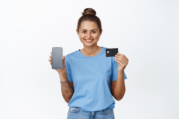 Clientes del banco. Una joven sonriente muestra su tarjeta de crédito y la pantalla de su teléfono inteligente, contra un fondo blanco.