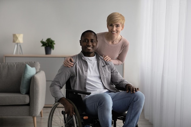 Clínica de reabilitação e atendimento ao paciente, enfermeira domiciliar ou família multirracial. Senhora caucasiana madura em marido afro-americano adulto casual e feliz em cadeira de rodas, na sala de estar ou no interior da clínica