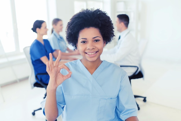 Clínica, profissão, pessoas e conceito de medicina - feliz médica afro-americana sobre grupo de médicos reunidos no hospital mostrando sinal de mão ok
