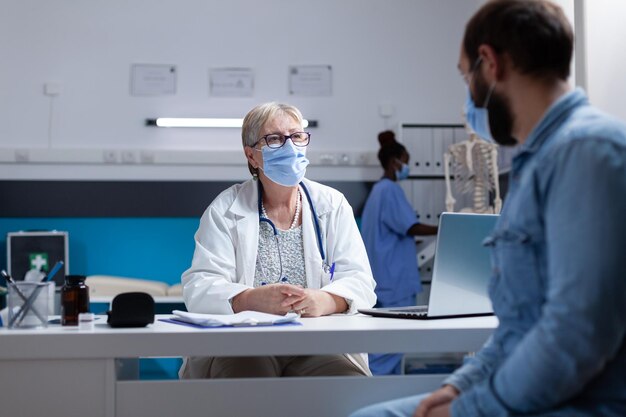 Clínico geral falando com o homem sobre doenças e saúde, usando máscara facial no gabinete. Paciente de consultoria médica no check-up médico durante a pandemia de coronavírus. Exame de saúde