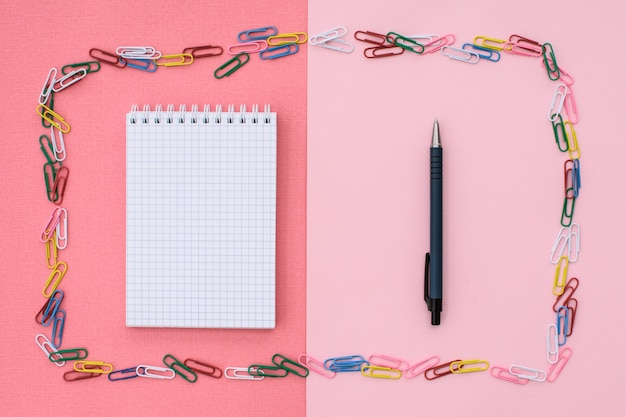 Clips de papel de colores en forma de marco y cuaderno en espirales en una jaula con bolígrafo sobre un fondo rosa coral. Concepto de regreso a la escuela. Lugar para el texto.