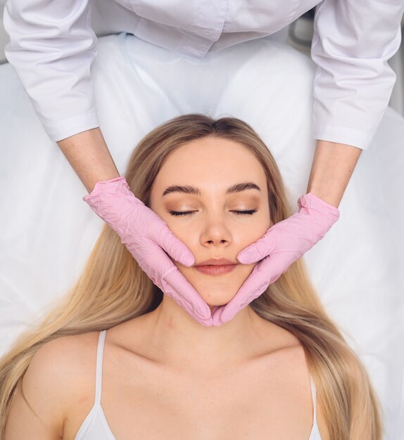 Close da esteticista com as mãos nas luvas, tocando o rosto de uma jovem mulher com cirurgia plástica facial