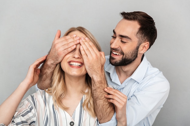 Close da foto de um homem bonito em roupas casuais sorrindo e cobrindo os olhos de sua namorada isolada sobre uma parede cinza