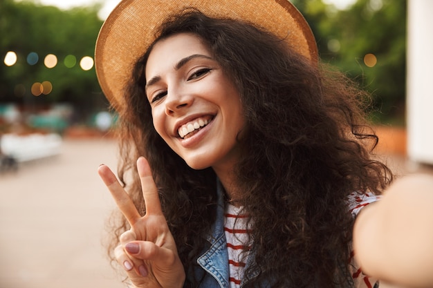 Close da foto de uma bela adolescente usando chapéu de palha, mostrando o símbolo da paz e tirando uma selfie enquanto caminha ao ar livre