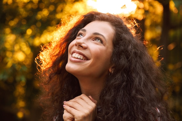 Close da foto de uma mulher morena sorridente, olhando para cima, enquanto caminha pelo parque em um dia ensolarado