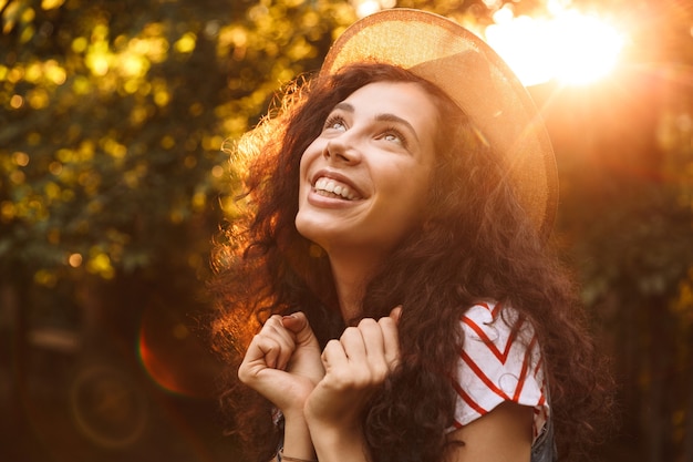 Close da foto de uma mulher morena sorridente usando chapéu de palha, olhando para cima, enquanto caminha pelo parque em um dia ensolarado