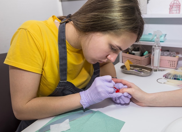 Close da garota do mestre cobrindo as unhas da cliente com verniz vermelho. uma manicure mestre trabalha em um salão de beleza. processo de hardware de manicure.