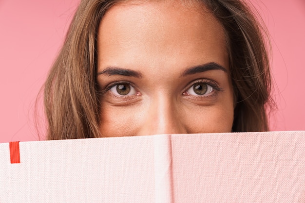 Close da imagem de uma linda garota sorrindo enquanto abraça seu livro de diário isolado sobre uma parede rosa