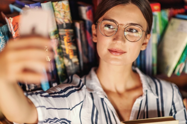 Foto close da jovem morena linda em pé na biblioteca, segurando um livro e tirando uma selfie.