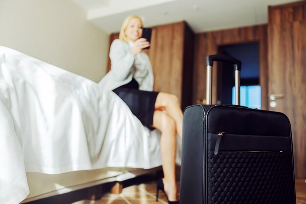 Close da mala no quarto do hotel. mulher de negócios sentada na cama e usando um telefone inteligente. ela está no simpósio.