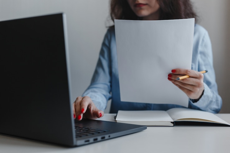 Foto close da mão de uma mulher com unhas vermelhas, segurando documentos e sentada na frente de um laptop de camisa azul.