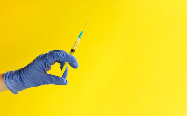 Foto close da mão do médico na luva azul, segurando a seringa de plástico sobre fundo amarelo.