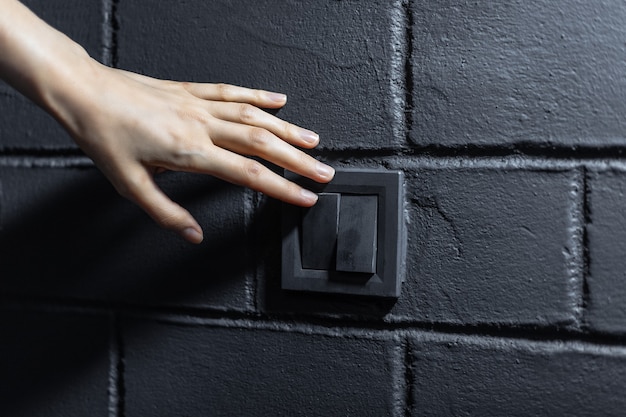 Close da mão feminina, acendendo / apagando a luz pelo botão do interruptor elétrico. Plano de fundo da parede de tijolos pretos.