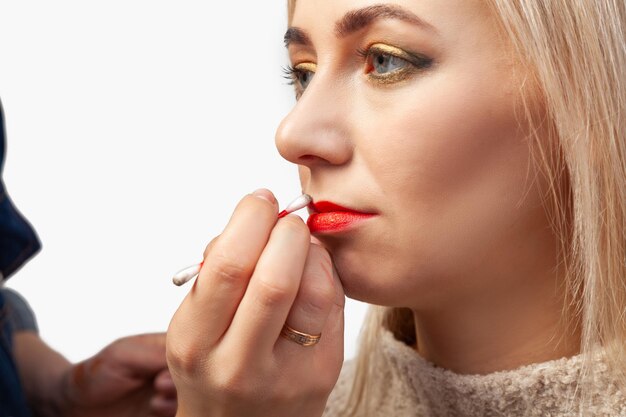 Close da maquiagem dos lábios de uma modelo com rosto claro o maquiador segura um cotonete nas mãos e lava o excesso de batom vermelho no contorno da boca