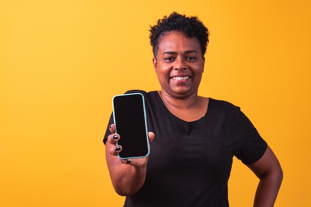 Close da mulher afro sorridente segurando o smartphone com uma tela preta em branco na mão em fundo amarelo.