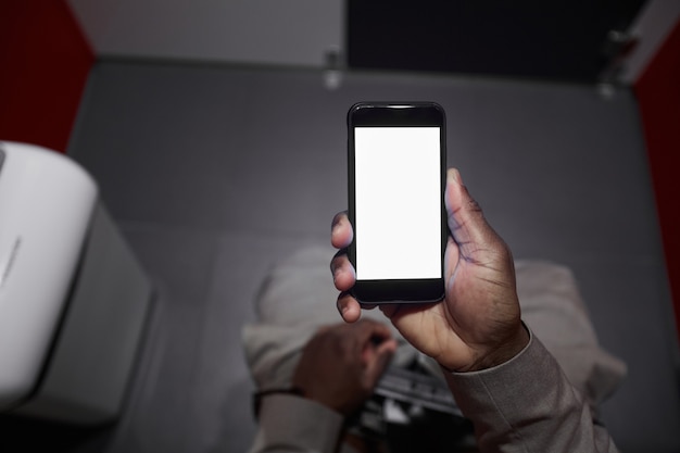 Close da vista superior de um homem afro-americano irreconhecível usando smartphone enquanto está sentado no banheiro em um banheiro público, copie o espaço