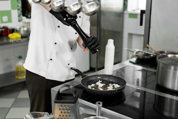 Foto close das mãos de um chef em uma túnica que adiciona sal ou pimenta a um prato ao fritar em uma frigideira.