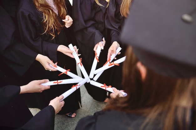 Close de estudantes de graduação em vestes pretas ou manto segurando diplomas ou certificados de graduação de uma instituição de ensino superior.