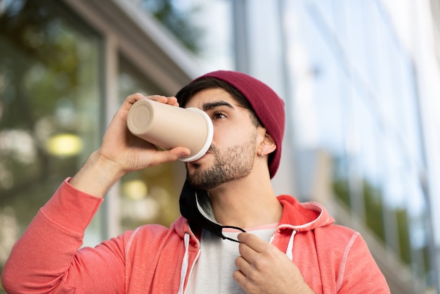 Close de jovem usando uma máscara protetora e bebendo café em pé ao ar livre na rua. Conceito urbano. Novo conceito de estilo de vida normal.