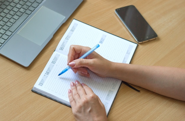 Close de mãos femininas escrevendo no caderno em uma mesa de madeira para smartphone e laptop ao lado