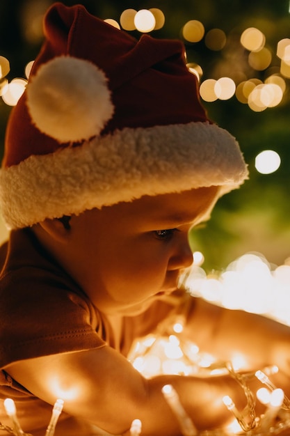 Close de um bebê com chapéu de papai noel, deitado de barriga no chão, em uma árvore de natal com luzes de fundo.