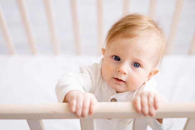 Close de um bebê loiro de 6 meses de idade sorridente em um berço em um quarto brilhante em um conceito de retrato de roupa de algodão branco de produtos infantis