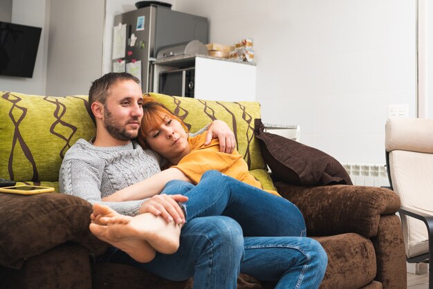 Foto close de um casal caucasiano se abraçando no sofá em casa assistindo televisão