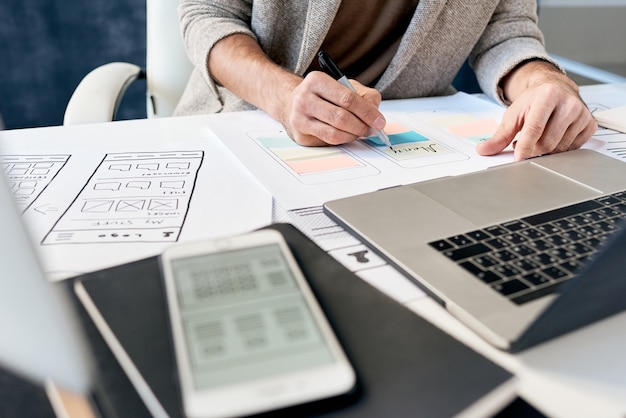 Close de um designer irreconhecível sentado à mesa com dispositivos e fazendo anotações sobre esquemas de design de telefone