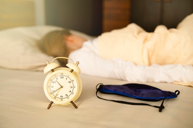 Foto close de um despertador e máscara de dormir, a menina dorme de costas. dorme por muito tempo, no final da manhã.